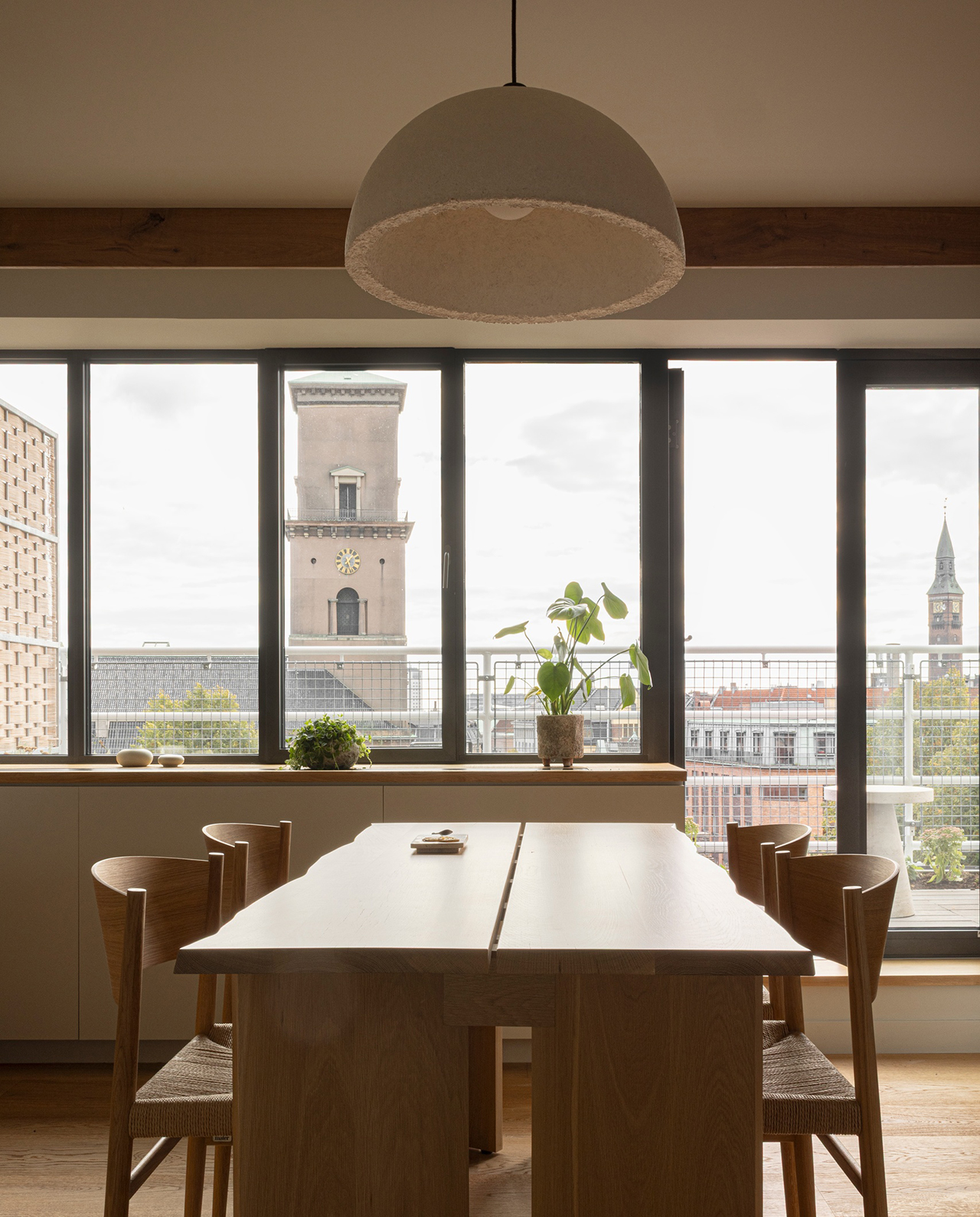 A modern dining room inspired by Norm Architects features a wooden table and chairs, large windows, potted plants, and a view of city buildings and clock towers outside.