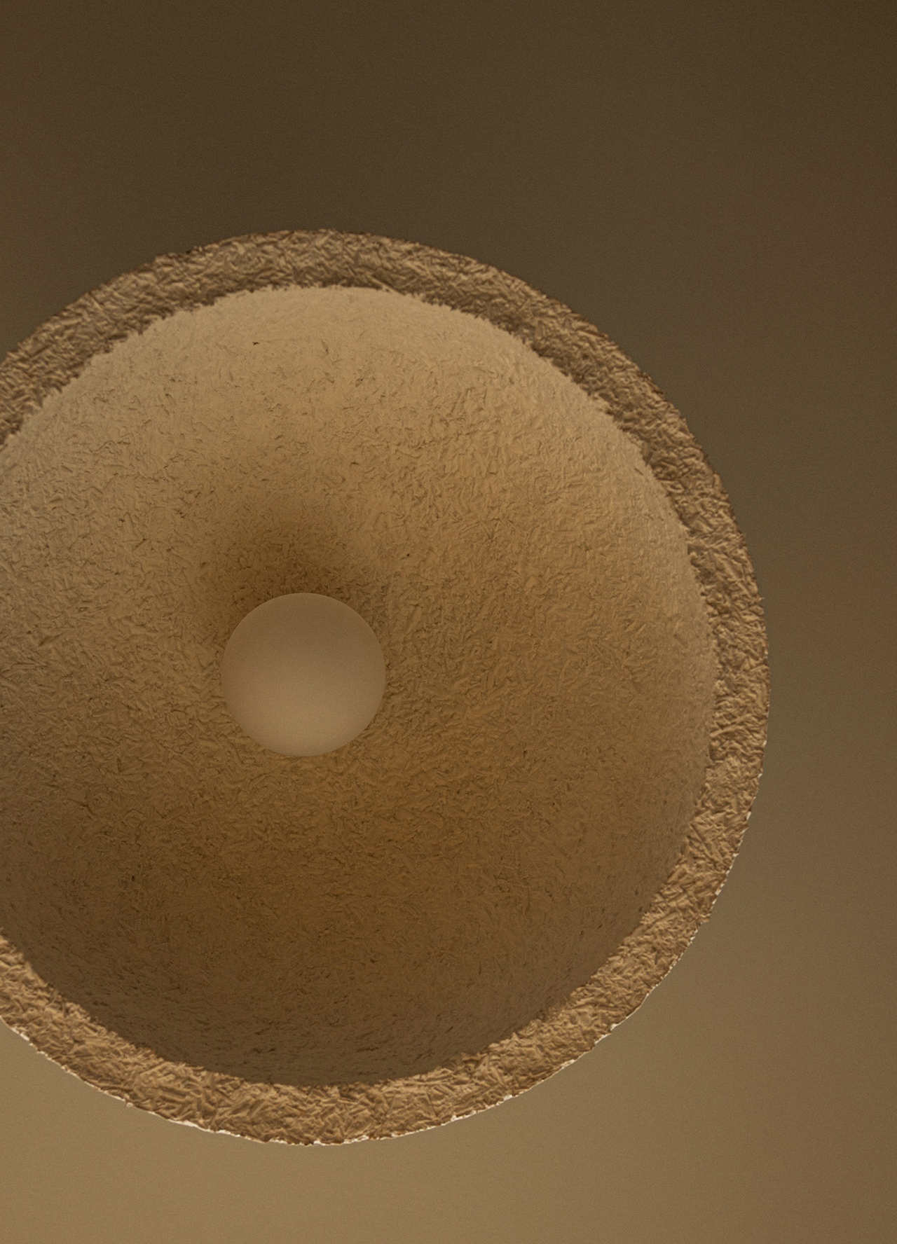 Close-up view of a textured, circular, beige lampshade designed by Norm Architects, with a lightbulb in the center, photographed from directly below against a brown background.
