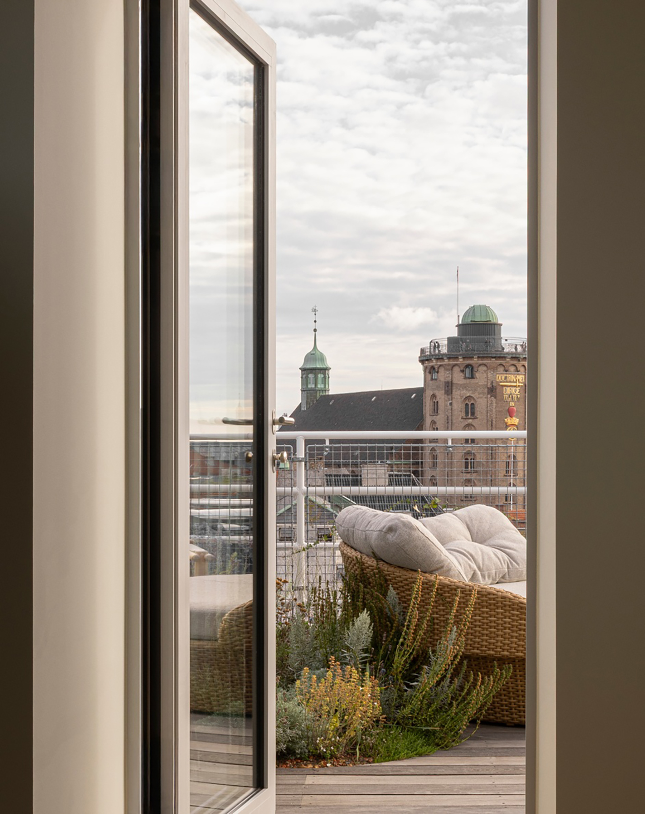 A glass door opens to a balcony with a cushioned wicker chair, railing, and plants, offering a city view with green domes in the background under a cloudy sky—a tranquil space inspired by Norm Architects.