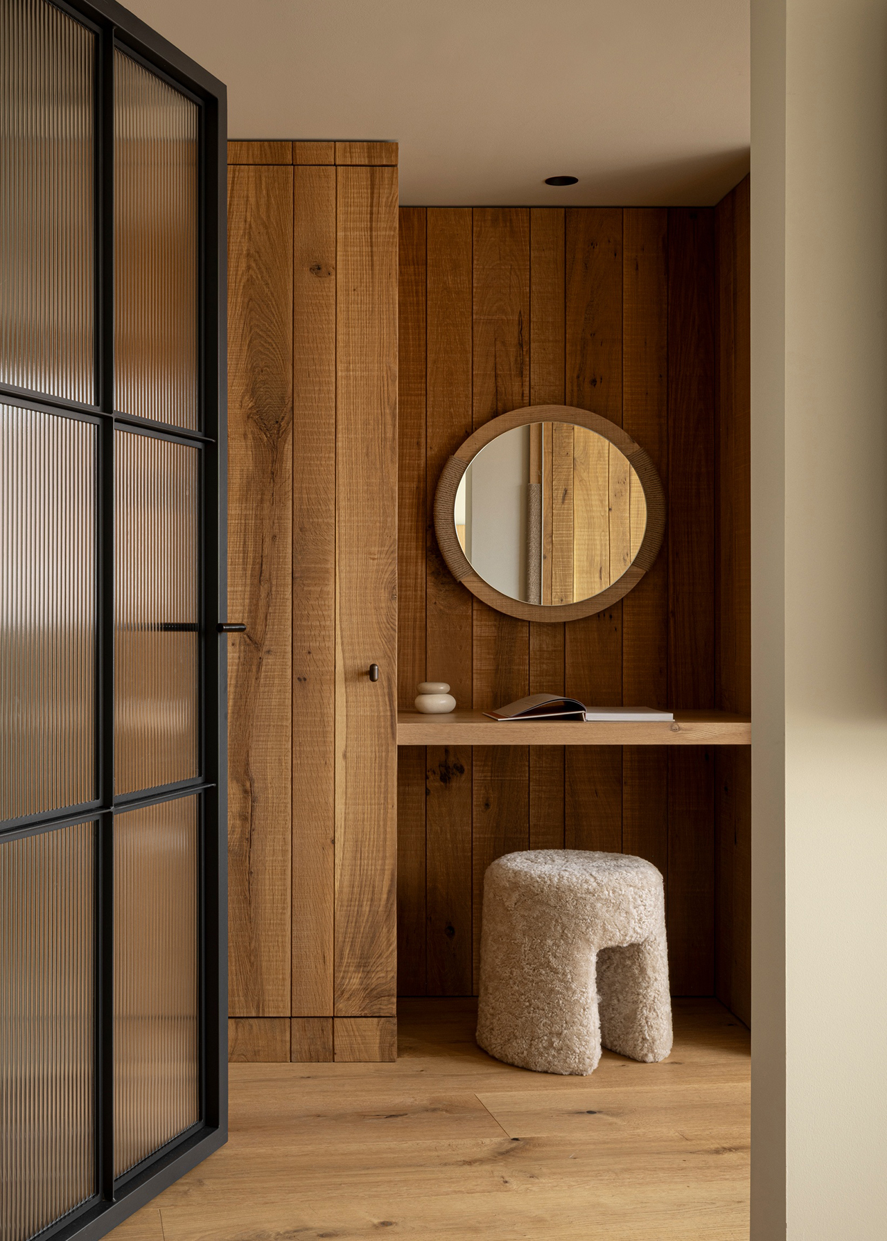 A small workspace by Norm Architects features a wooden desk, round mirror, textured stool, and wood-paneled walls, viewed through a glass door. An open book and small decor items rest on the minimalist desk.