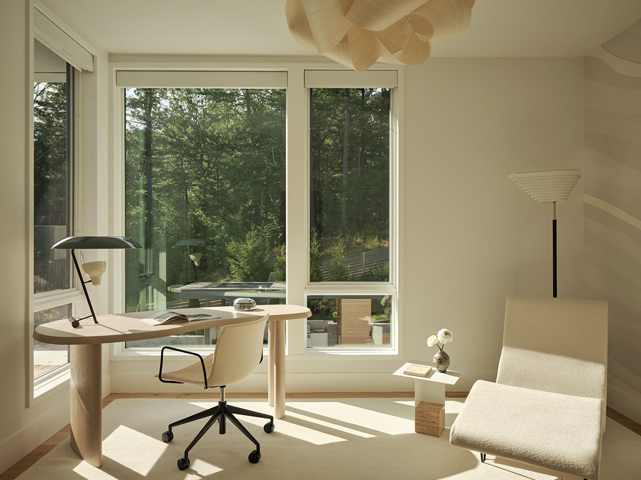 Minimalist home office with a curved wooden desk, white office chair, lounge chair, small side table, and large windows overlooking a green outdoor view.