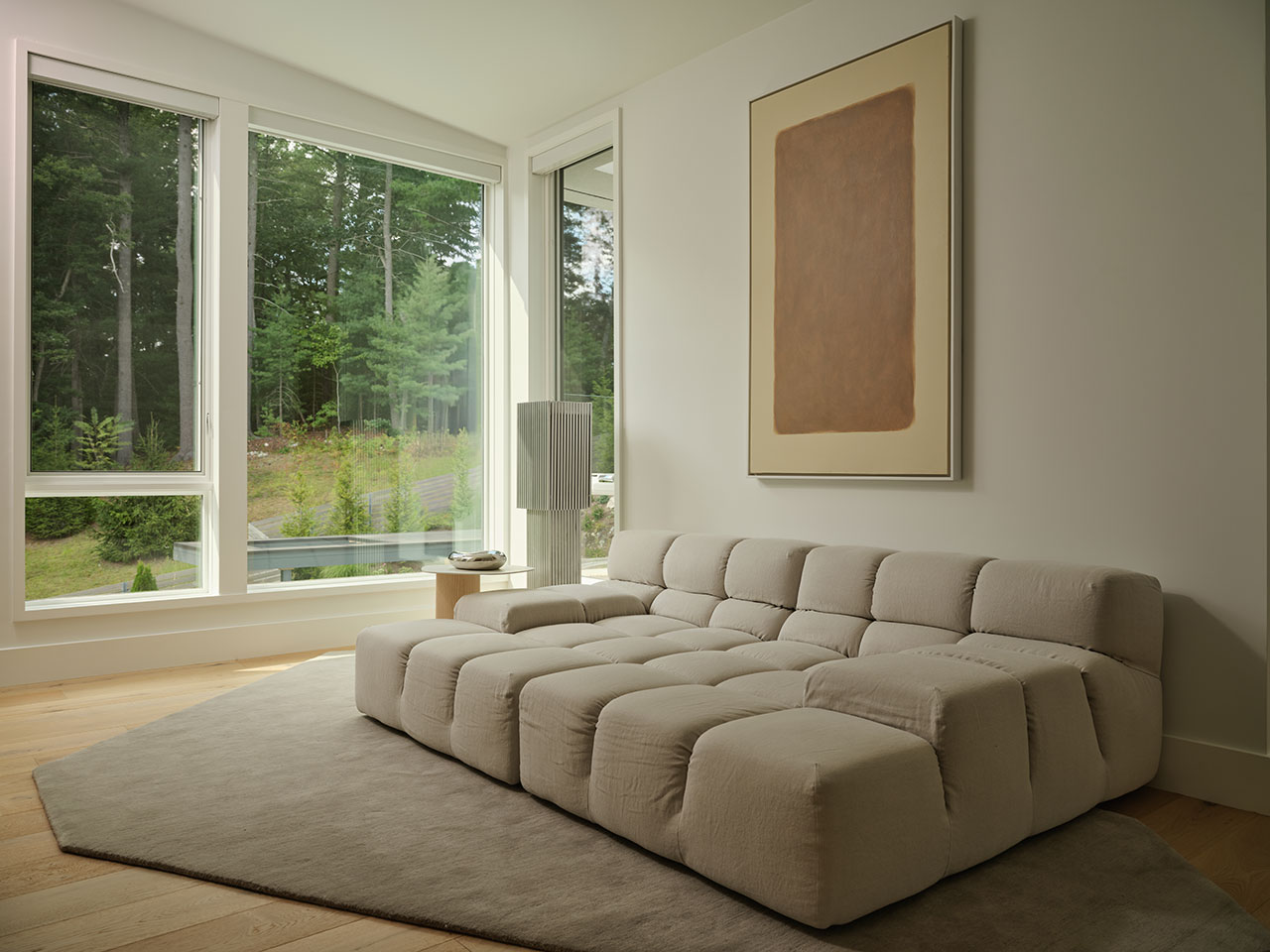 A modern living room with a large beige sectional sofa, a neutral rug, a side table with a lamp, a minimalist abstract painting, and large windows overlooking a green outdoor area.
