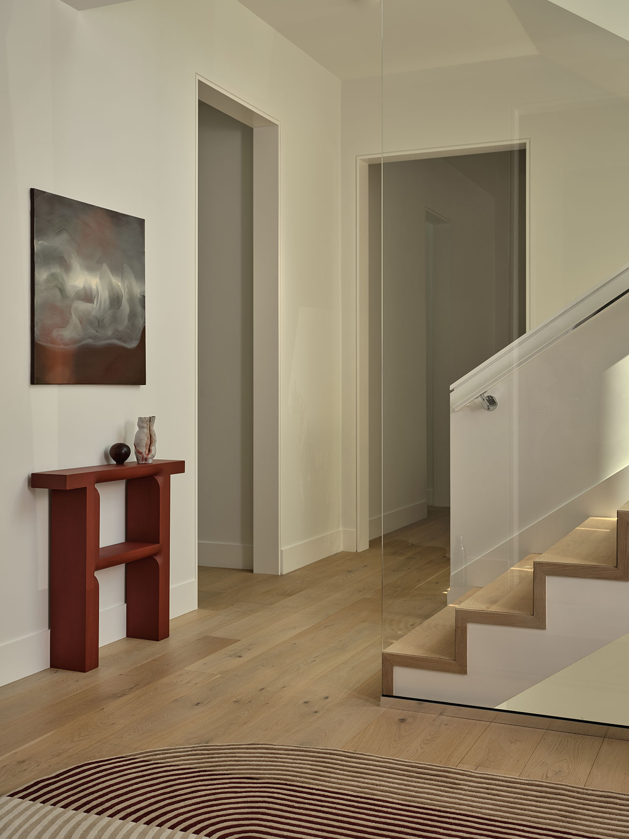 Modern interior hallway with light wood flooring, white walls, a staircase with glass railing, a red console table, and abstract wall art.