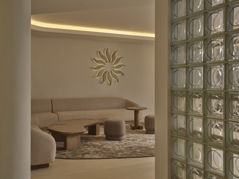 Minimalist living room with beige curved sofa, round poufs, wooden tables, abstract sun wall art, and a glass block partition. Neutral tones and soft lighting create a calm atmosphere.