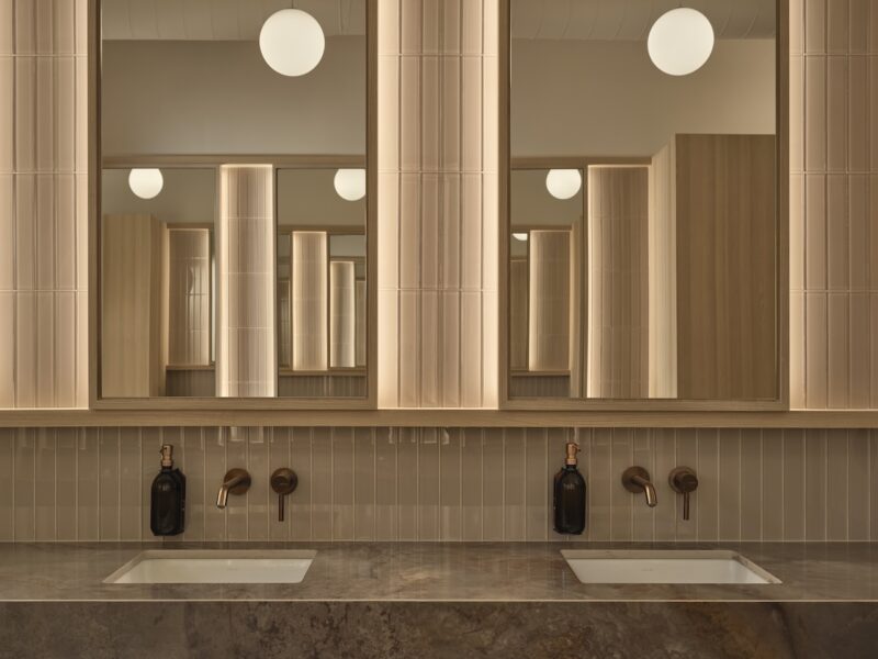 Modern public restroom with two sinks, wall-mounted soap dispensers, large mirrors, beige tiles, and globe pendant lights reflected in the mirrors.