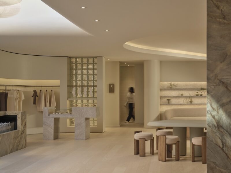 Minimalist, beige-toned interior with marble furniture, clothing racks, shelves, and a person walking in the background.