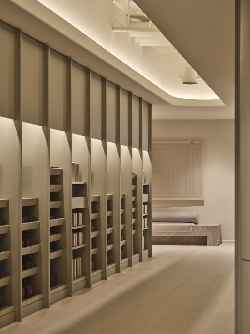 Minimalist, softly lit interior with floor-to-ceiling shelves holding books and objects, light wood flooring, and a cushioned bench against the wall.