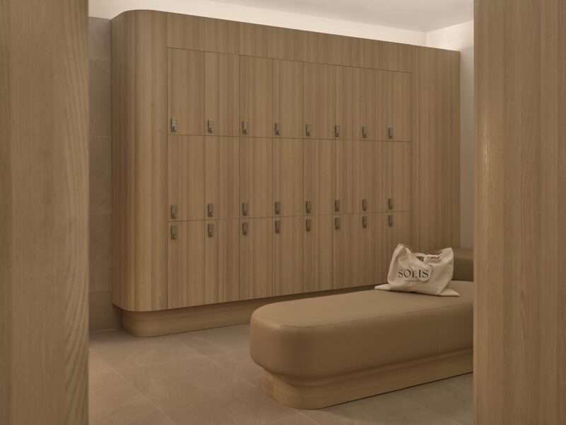 A locker room with light wood lockers, a beige cushioned bench, and a tote bag labeled