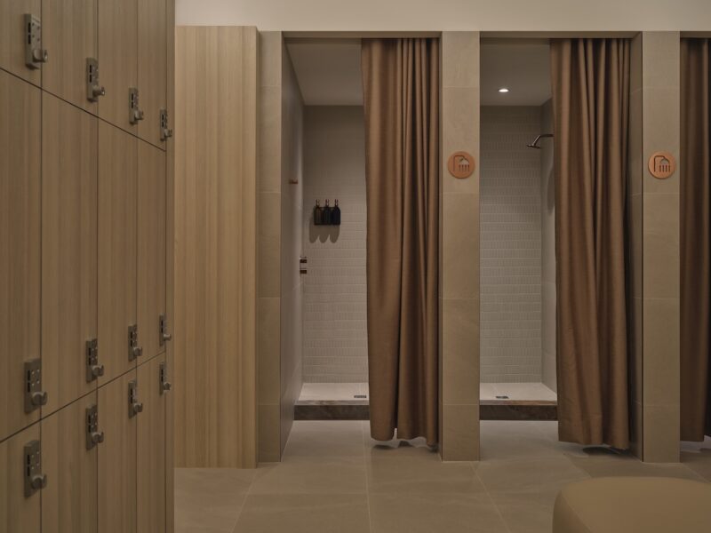 A modern locker room with beige lockers on the left and two shower stalls with brown curtains and wall-mounted toiletries on the right.