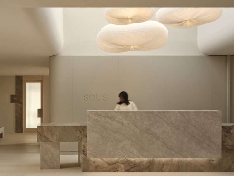 A person stands behind a marble reception desk in a modern, minimalist lobby with soft lighting and sculptural ceiling lights.