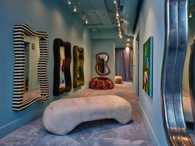 A hallway with blue walls, three wavy-framed mirrors, a curved beige bench, a brown pouf, carpeted floor, and wall art under track lighting.