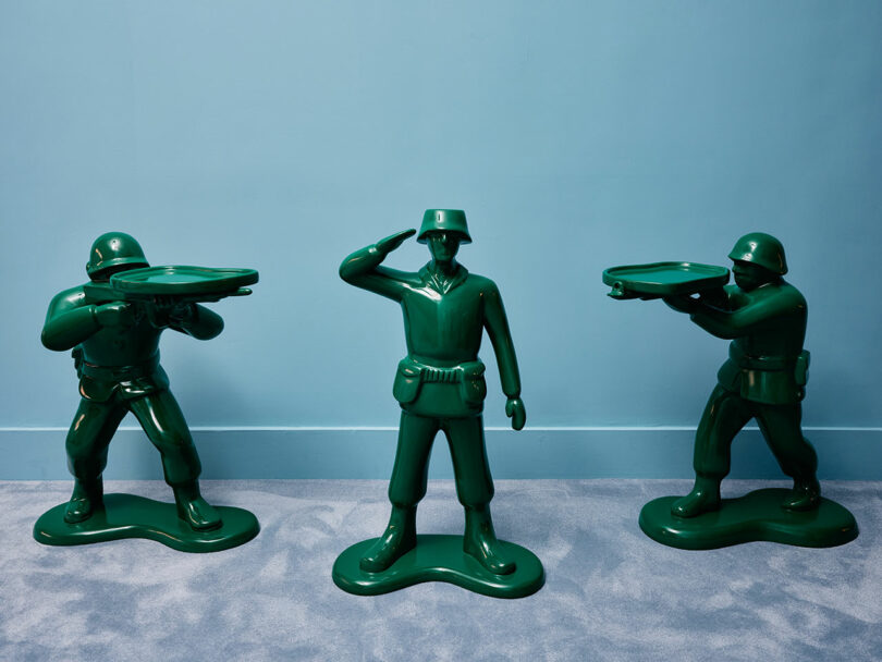 Three oversized green plastic toy soldiers stand on a blue floor against a blue wall, each holding a tray as if serving or presenting something.