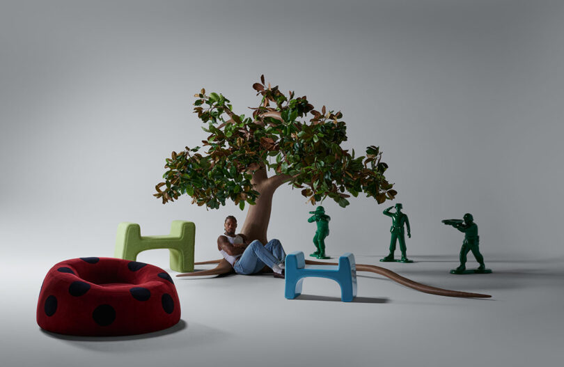 A person sits under a large artificial tree, surrounded by oversized green toy soldiers, a red polka-dotted seat, and two colorful abstract stools in a minimal studio setting.