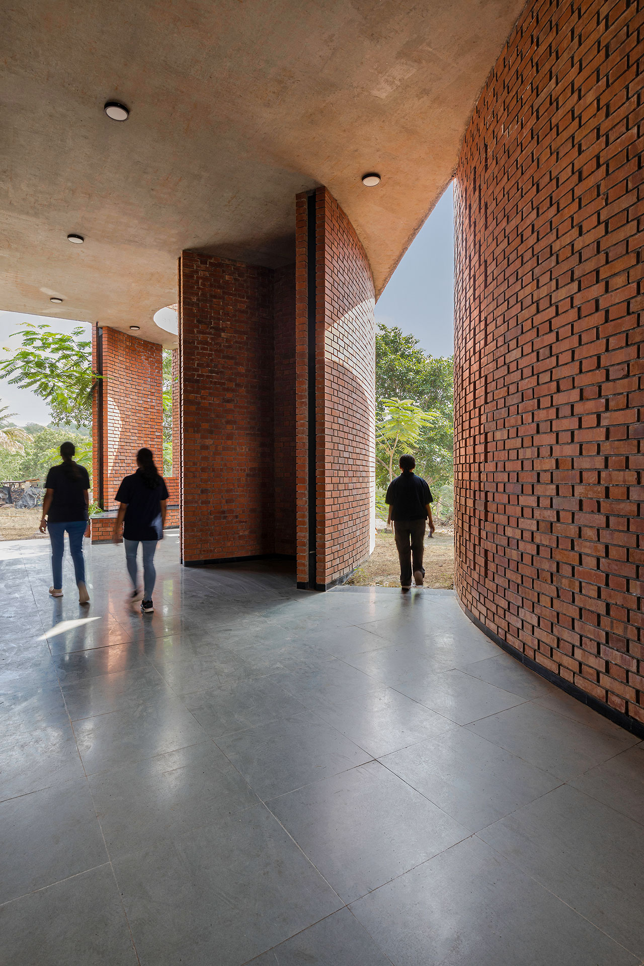 Three people walk through a modern building corridor shaped by morphogenesis, with tall curved brick walls and large window openings letting in natural light.