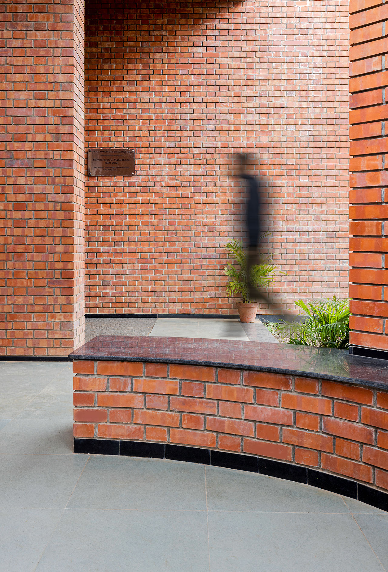 A blurred person walks past red brick walls and a potted plant, illustrating the morphogenesis of movement in an outdoor corridor with grey flooring.