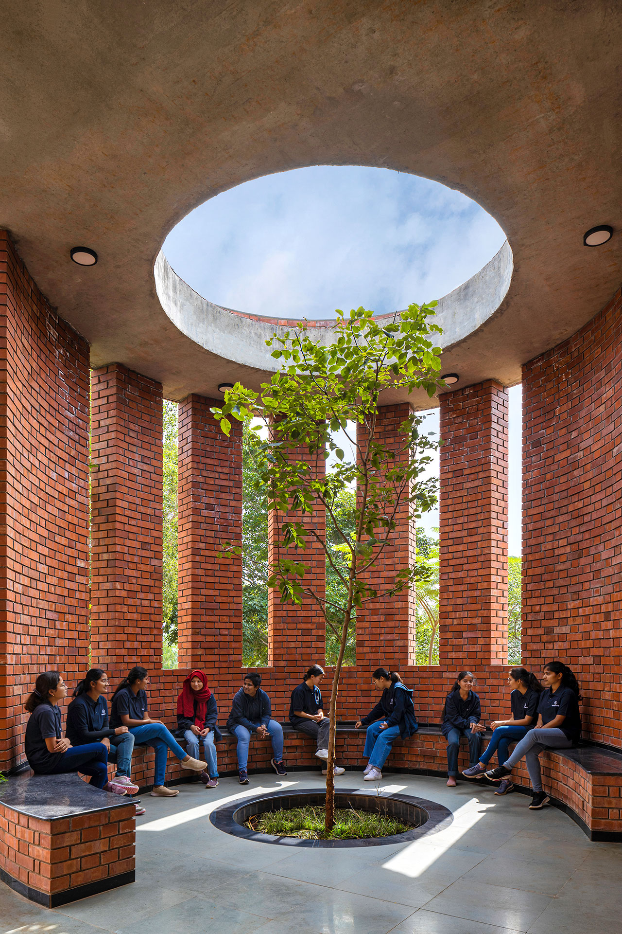 A group of people sits in a circular brick pavilion, its design inspired by morphogenesis, with a central tree and an open skylight above that allows natural light to fill the space.