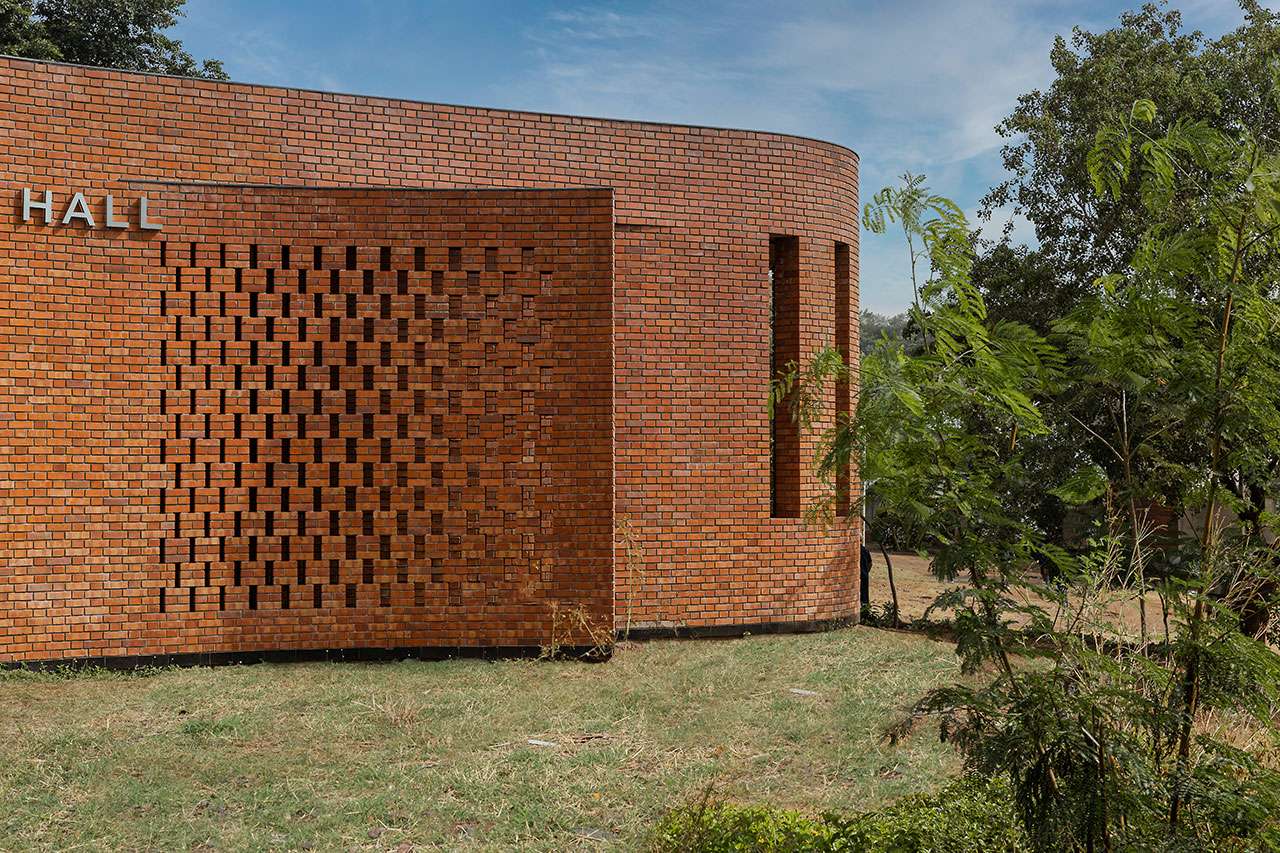 Curved brick building with patterned brickwork and narrow vertical windows showcases morphogenesis in architecture, partially surrounded by grass and trees under a blue sky.