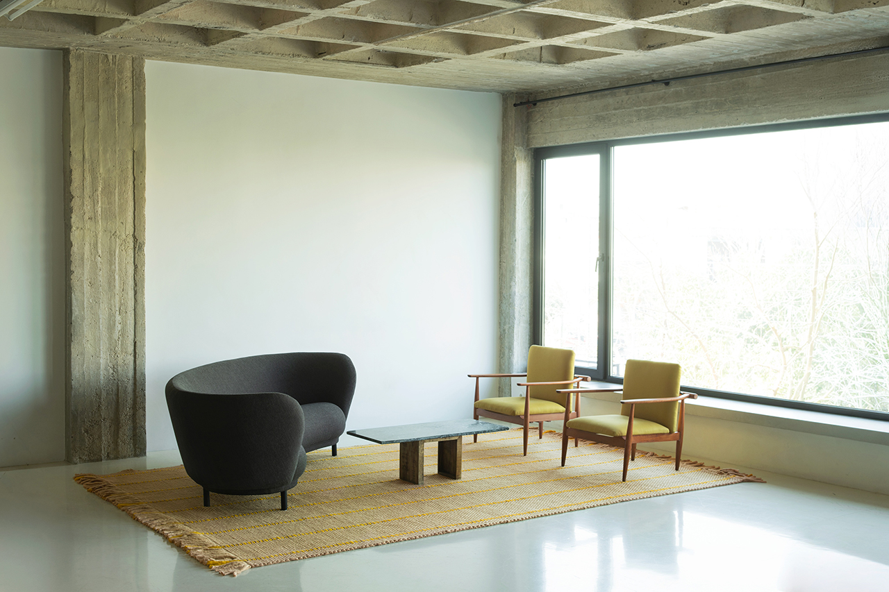 A minimalist room with a concrete ceiling, large window, a dark curved sofa, two yellow armchairs, a rectangular coffee table, and a woven rug creates a LAYERED look with its thoughtful mix of textures and forms.