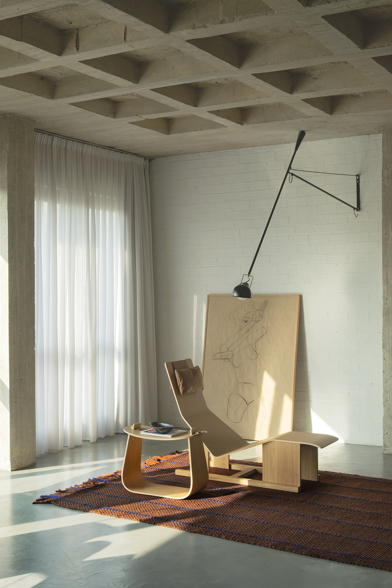 A minimalist room with a modern wooden lounge chair on a layered, textured rug, a large sketch leaning against a white brick wall, and a wall-mounted lamp above. Light filters through sheer curtains.