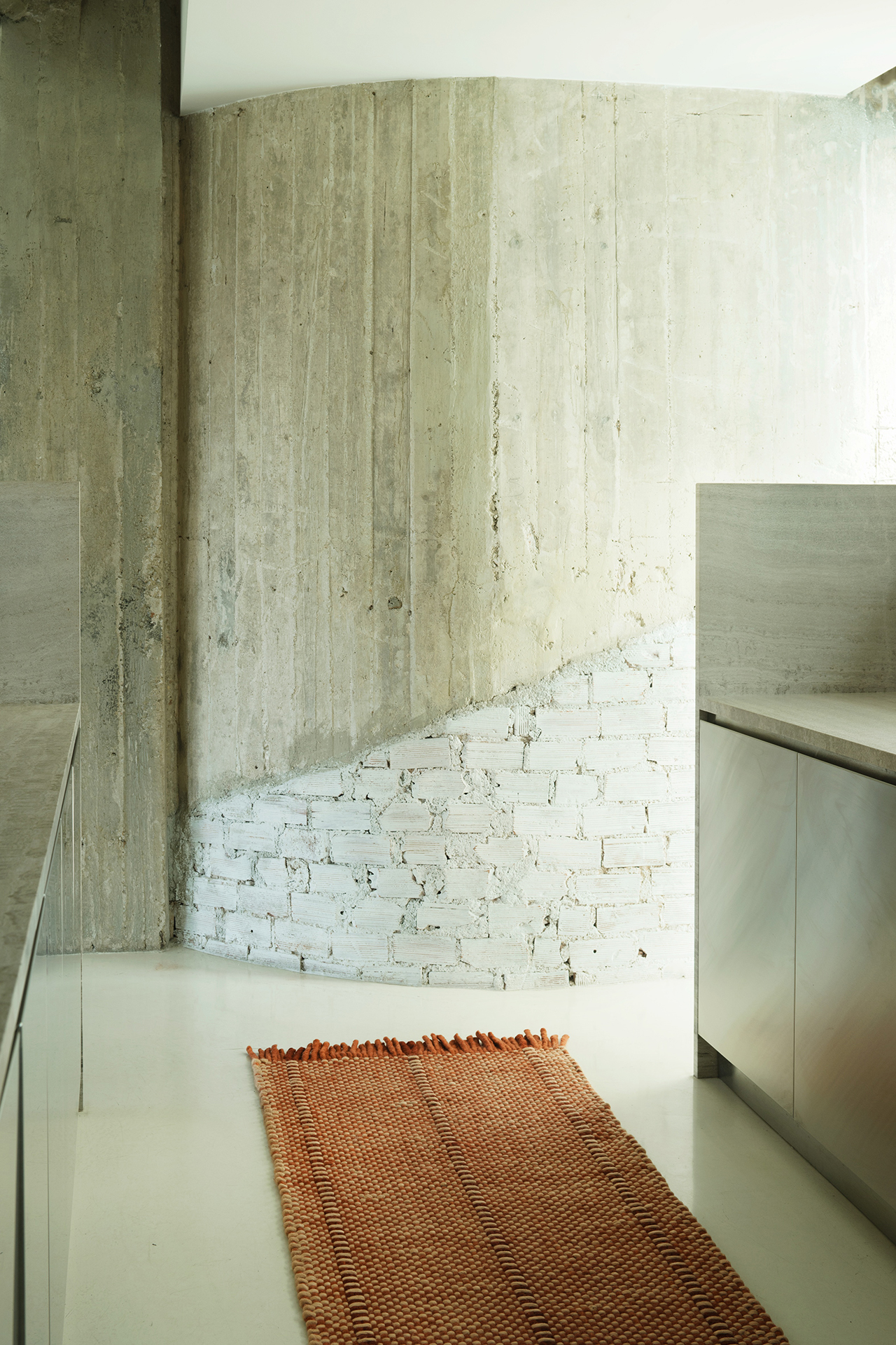 A minimalist kitchen with LAYERED concrete and white brick walls, stainless steel cabinets, and an orange woven rug on a light floor.
