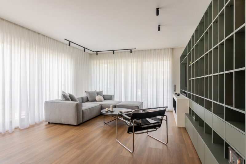 Modern living room with large windows, sheer white curtains, a gray sectional sofa, a black chair, a coffee table, and a green built-in shelving unit on the right wall.