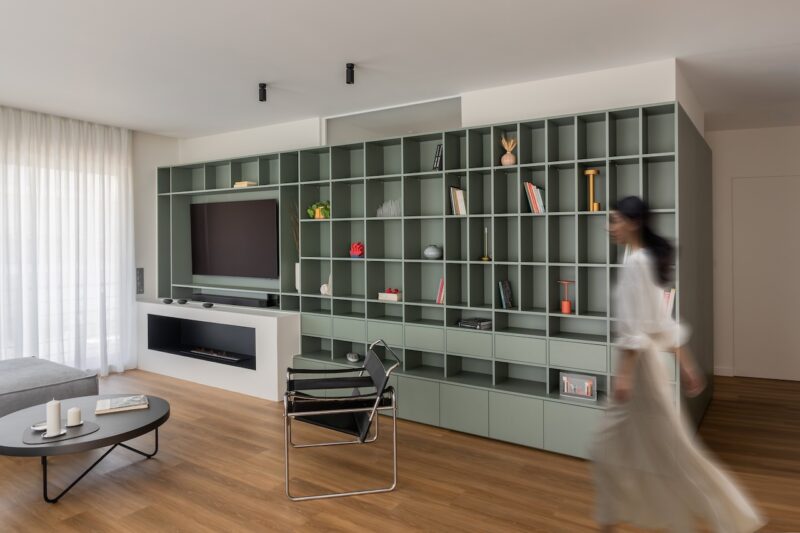 A modern living room with green built-in shelving, a TV, fireplace, minimalist decor, and a blurred person walking past a black chair on wood flooring.