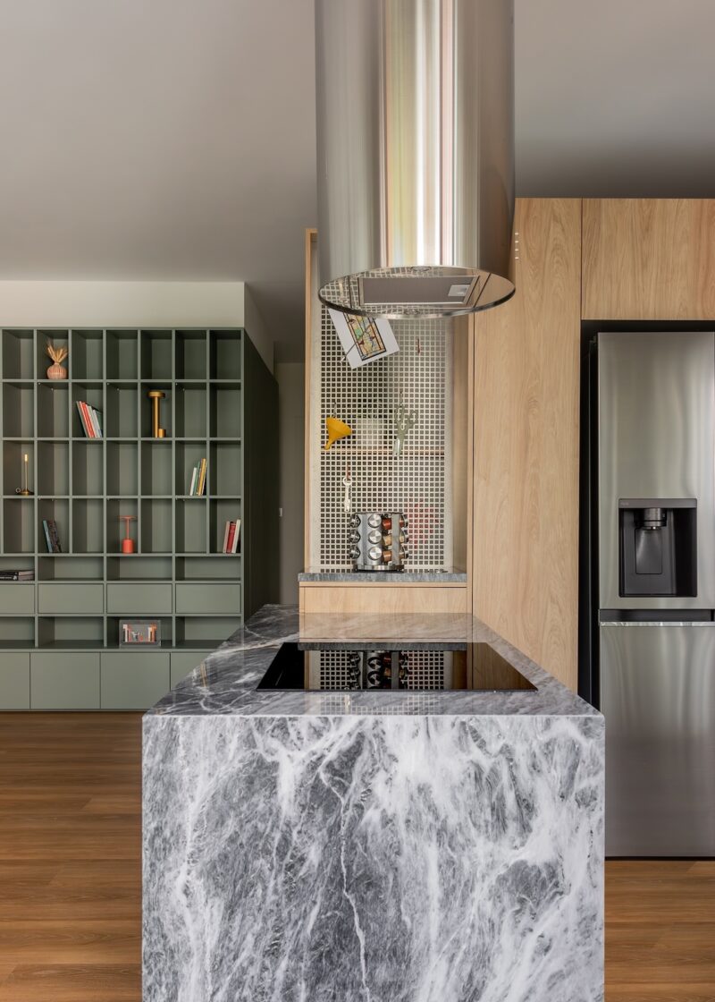Modern kitchen with a marble island, built-in stovetop, stainless steel refrigerator, wooden cabinets, and open shelving with books and decor in the background.
