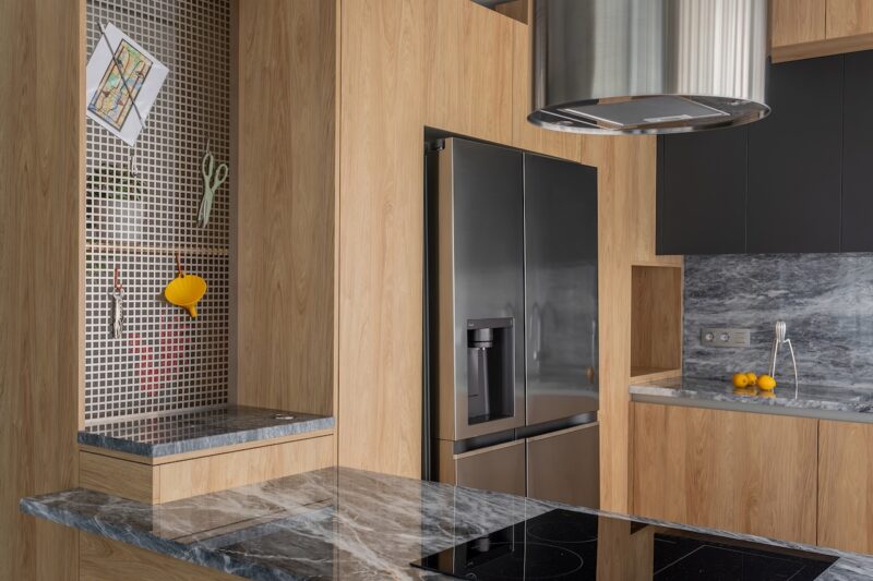 Modern kitchen with wood paneling, gray marble countertops, a black refrigerator, and a pegboard holding scissors, a postcard, and a lemon juicer.
