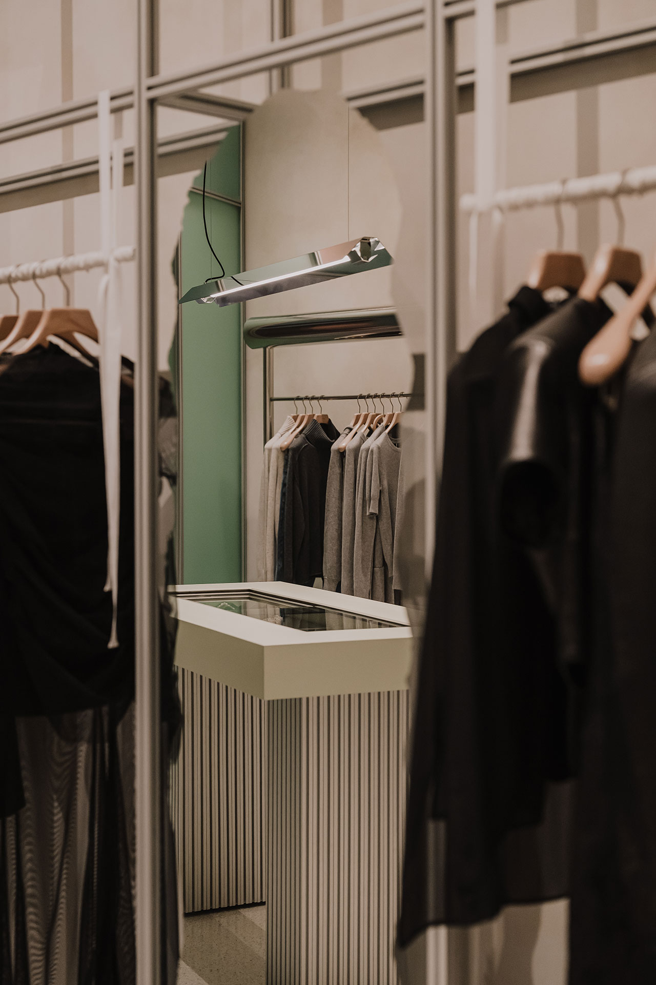 Clothing racks with black and gray garments are reflected in a mirror next to a modern counter in a minimalist retail store.