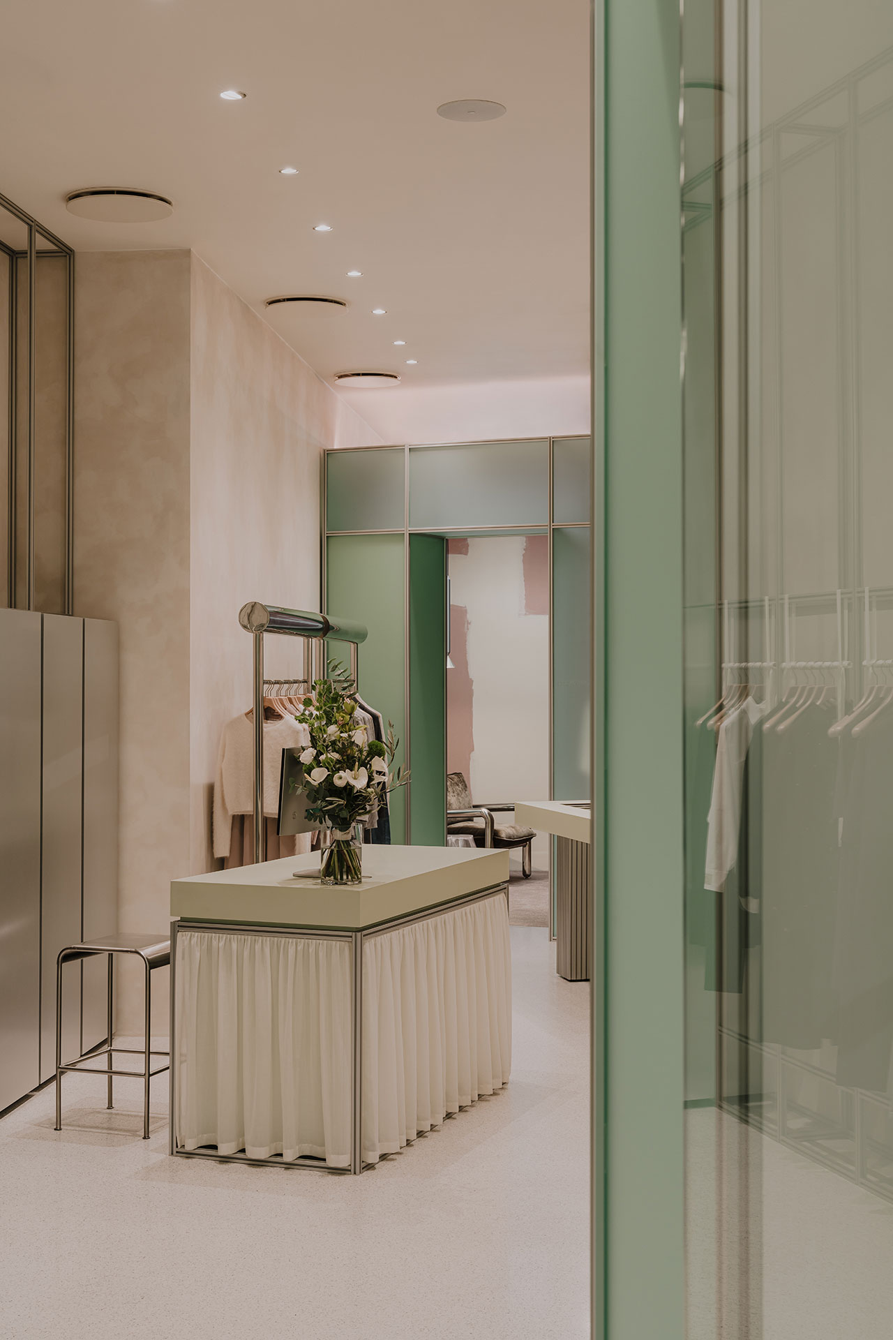 A modern boutique interior with soft lighting, pastel green accents, a central display table with a flower arrangement, and clothing racks along the walls.