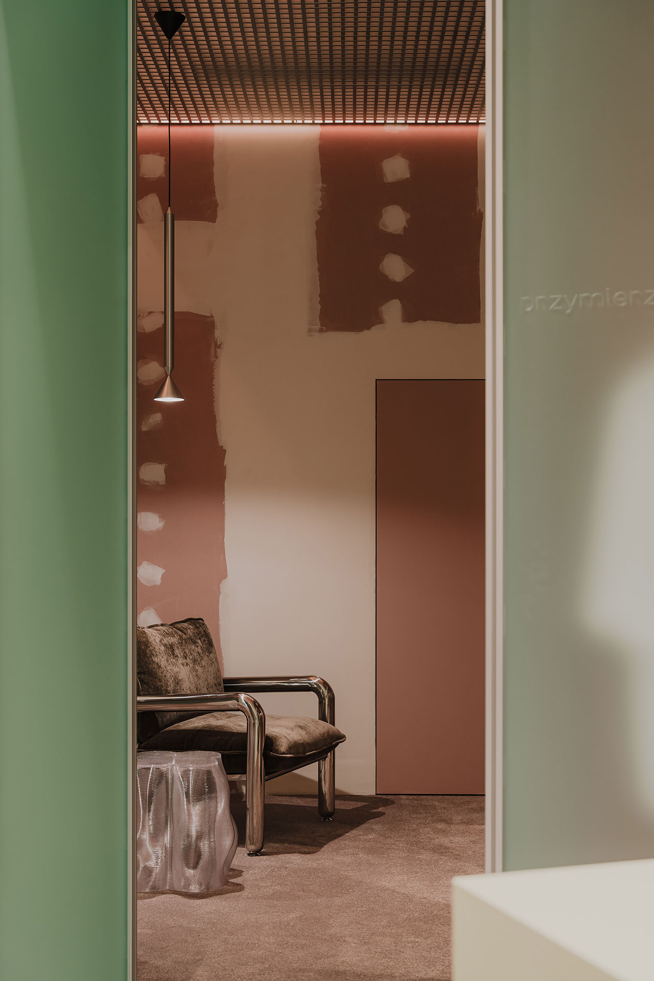 A minimal room with unfinished pink and white walls, a metal-framed chair with cushions, a clear side table, and a hanging pendant light.
