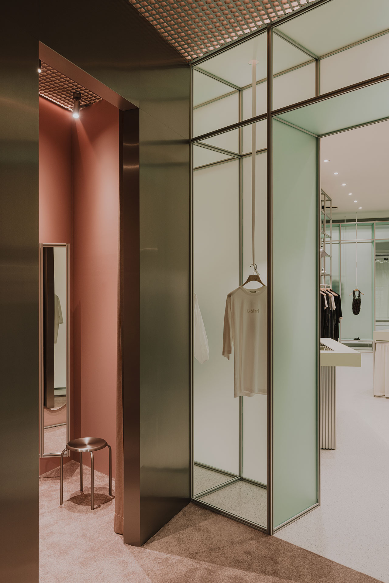 Modern fitting room with a full-length mirror, metal stool, and glass walls; a white t-shirt hangs on a rack inside the adjacent area.
