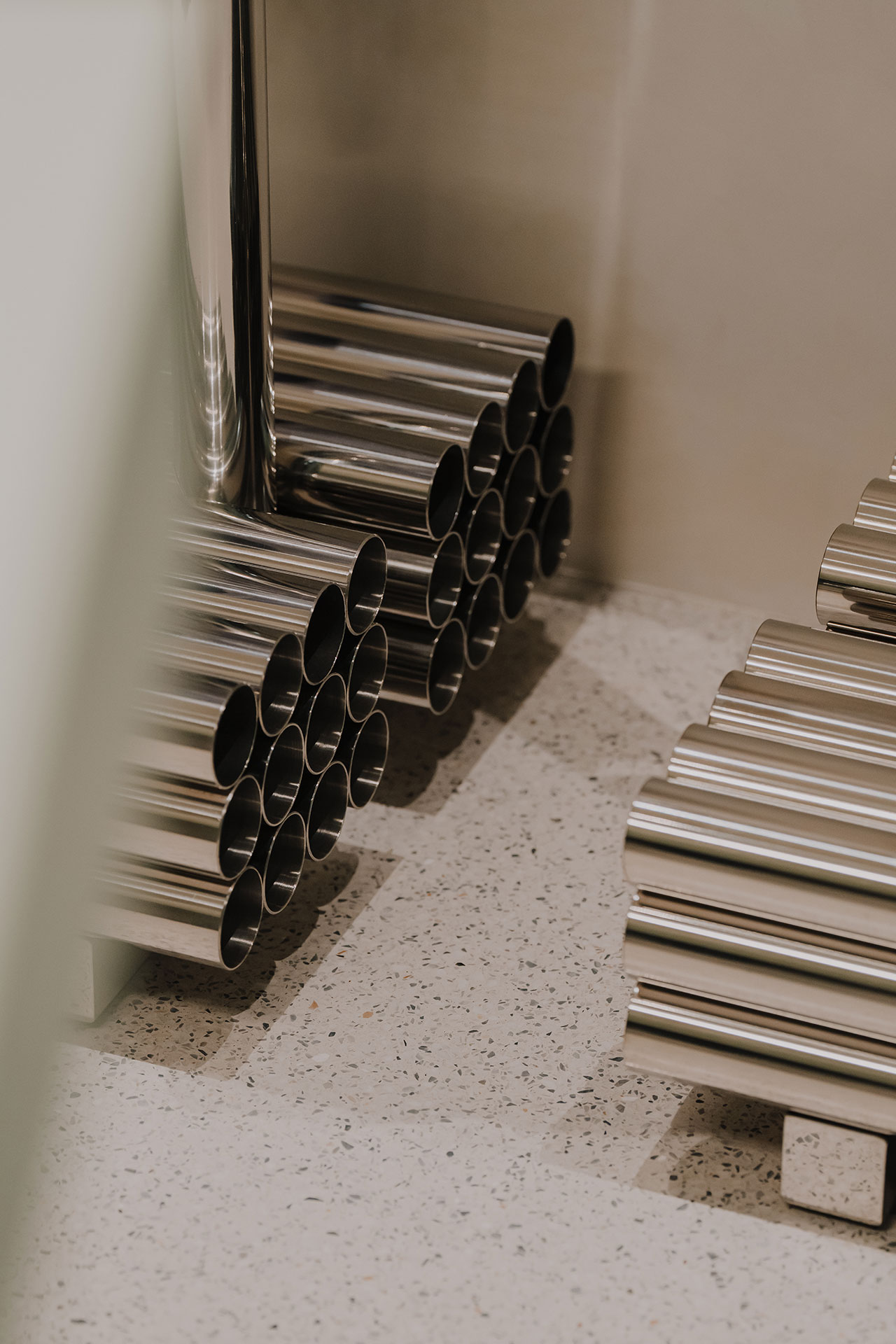 Several stacks of shiny metal pipes arranged parallel to each other on a speckled floor against a beige wall.