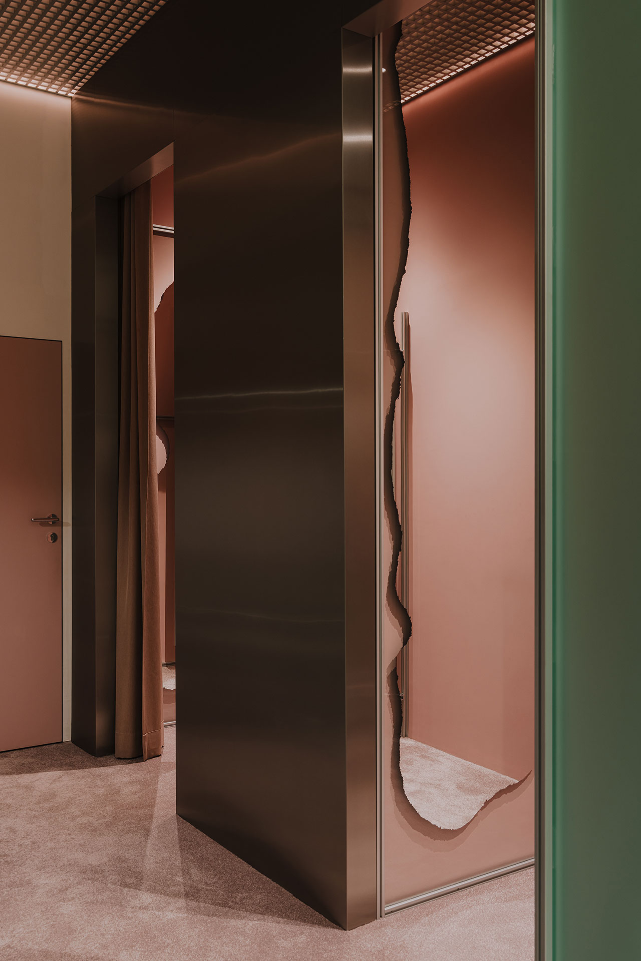 A modern interior features a metallic doorway with an irregular, jagged-edged mirror beside a muted pink door and walls, and soft beige carpet flooring.