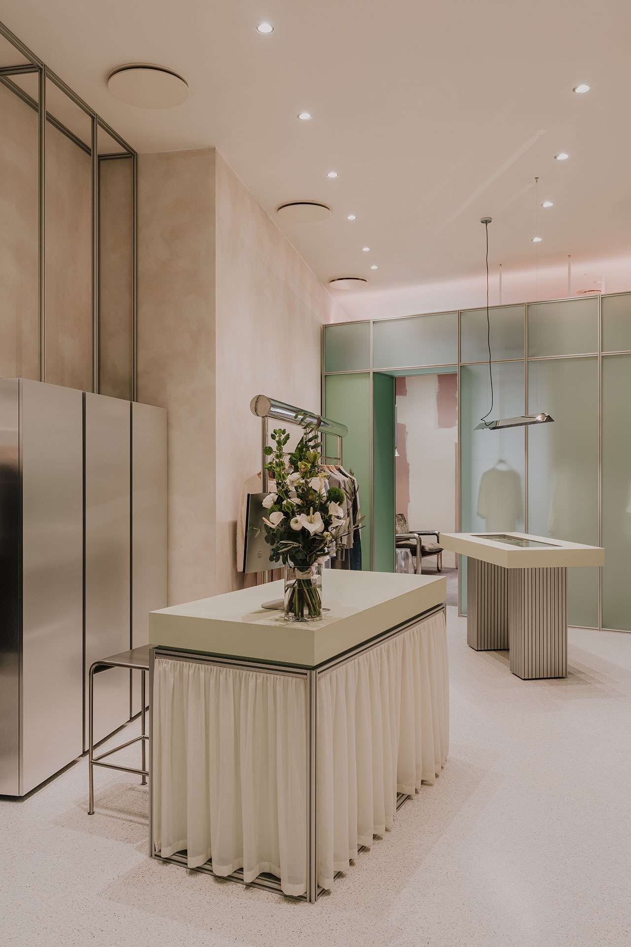 A modern, minimalist interior features a central table with a vase of flowers, stainless steel cabinets, and soft lighting, with sheer white fabric draped around the table’s base.