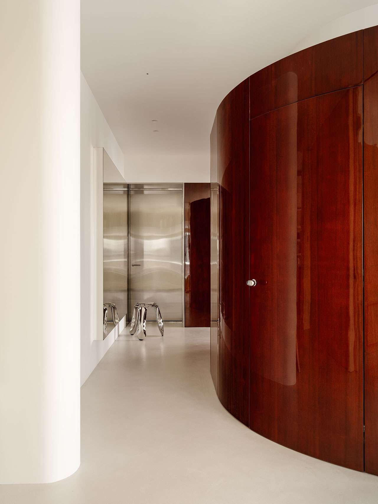 Minimalist hallway with a polished wood curved wall, white floor and walls, a stainless steel door, and an Antwe reflective metallic stool.