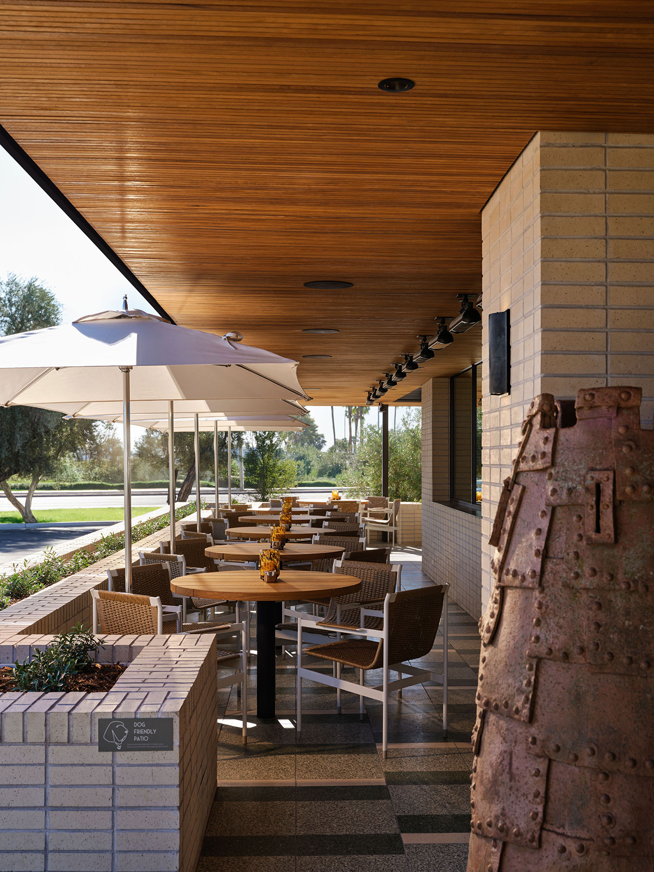 Outdoor restaurant seating area with wooden ceiling, cream brick walls, tables set with yellow flowers, wicker chairs, and large white umbrellas. A sculptural metal figure stands in the foreground.