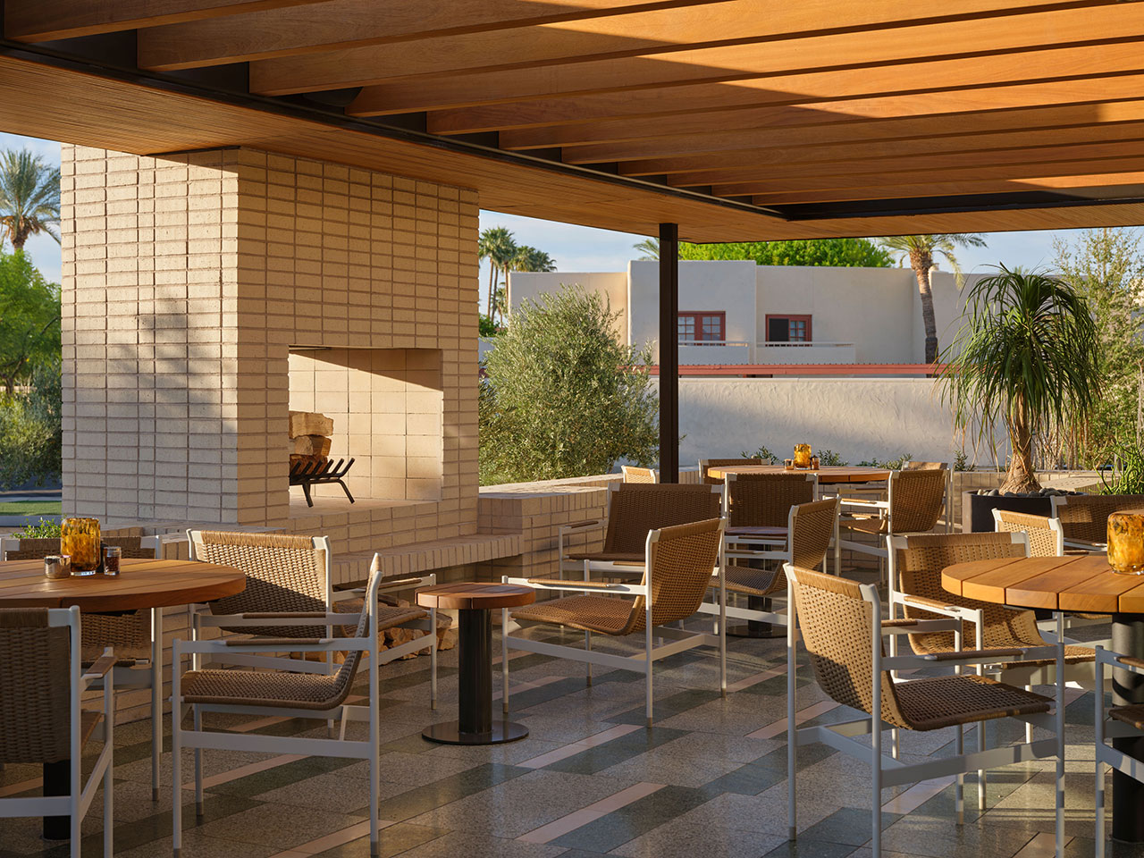 Outdoor patio with wooden ceiling, wicker chairs, round tables, a brick fireplace, and potted plants, set in a sunny, modern courtyard.