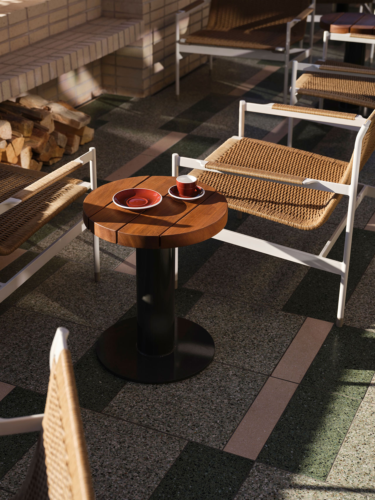 Small round wooden table with two red cups and saucers, surrounded by woven chairs on a tiled floor in an outdoor seating area.