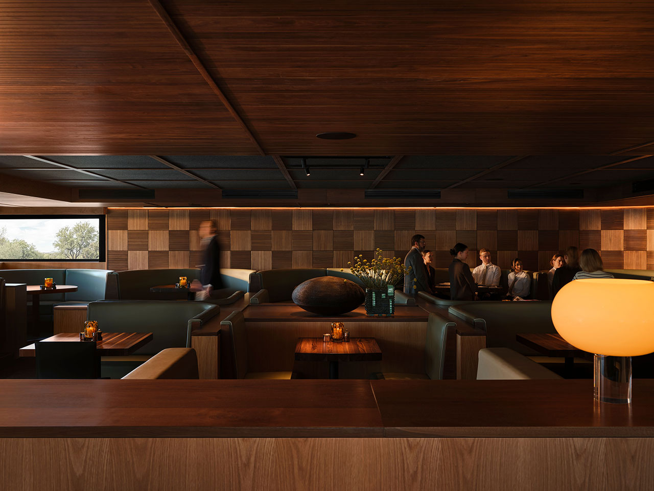 A modern restaurant interior with wood-paneled walls, leather booths, dim lighting, and a group of people conversing in the background.
