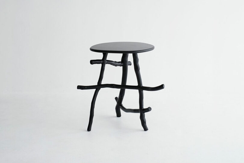 A small, round, black side table with three uneven, branch-like legs and additional crossbars, set against a plain white background.
