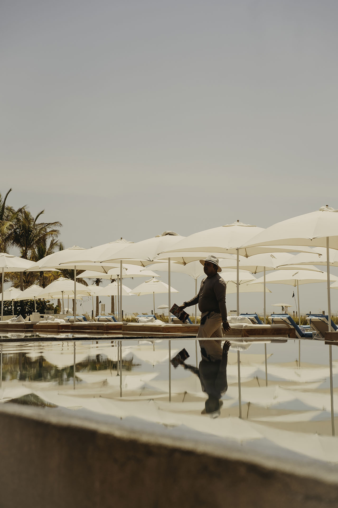 A person carrying a tray walks by a poolside area with white umbrellas and lounge chairs under a clear sky.