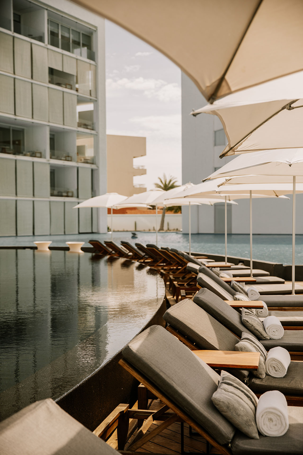 Poolside loungers with rolled towels and umbrellas beside a modern hotel building, overlooking a calm outdoor swimming pool.