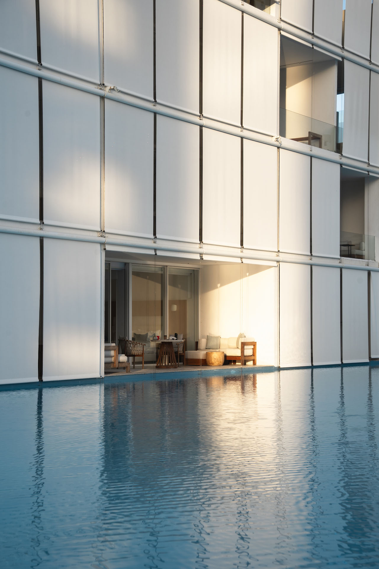 Modern building facade with large white panels, an outdoor seating area on a wooden deck, and a calm swimming pool in the foreground.