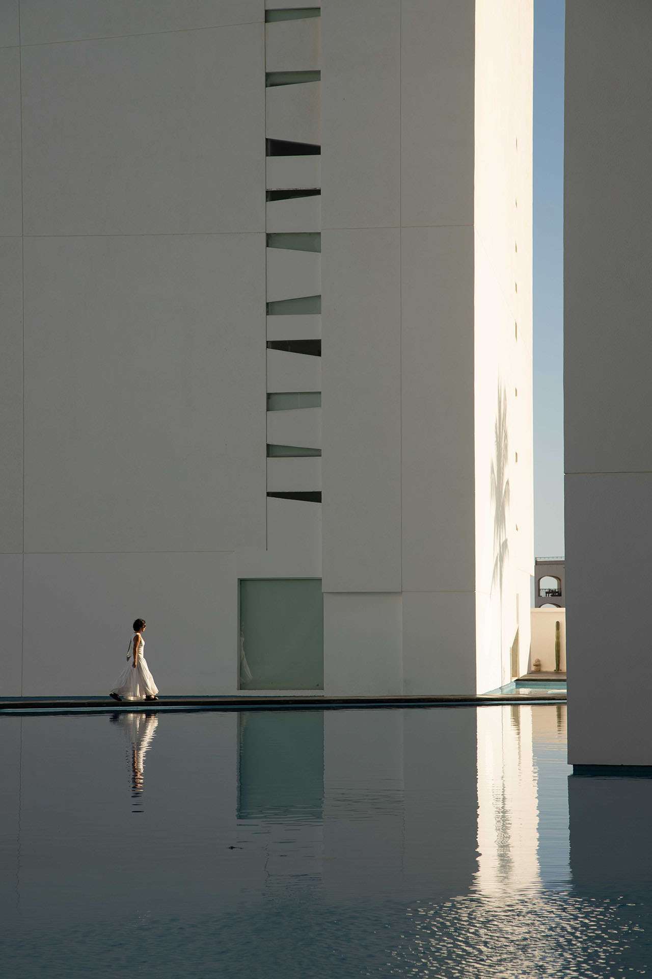 A person in a white dress walks alongside a modern, white building reflected in a still pool of water under clear daylight.