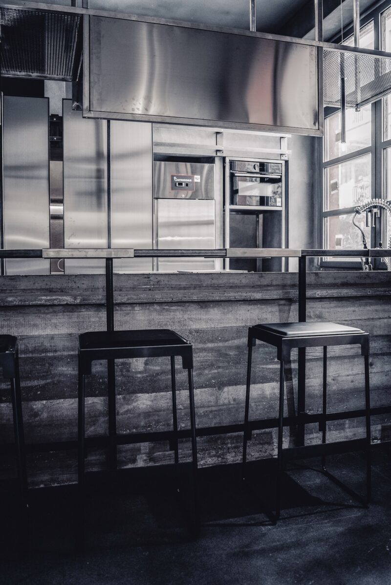 Modern industrial kitchen with a metallic finish, featuring a bar counter and three black bar stools in front of stainless steel appliances and large windows.