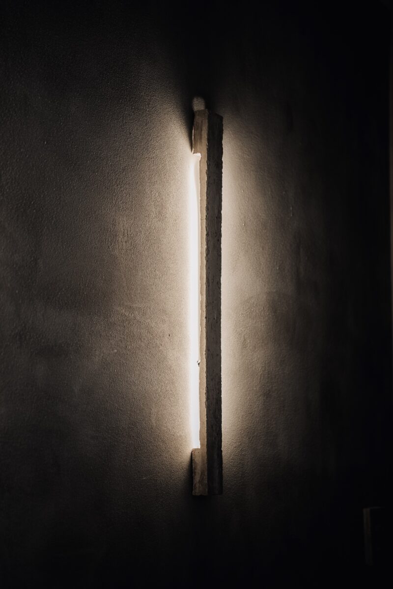 Vertical wall-mounted light fixture emits a warm glow against a dark, textured wall.