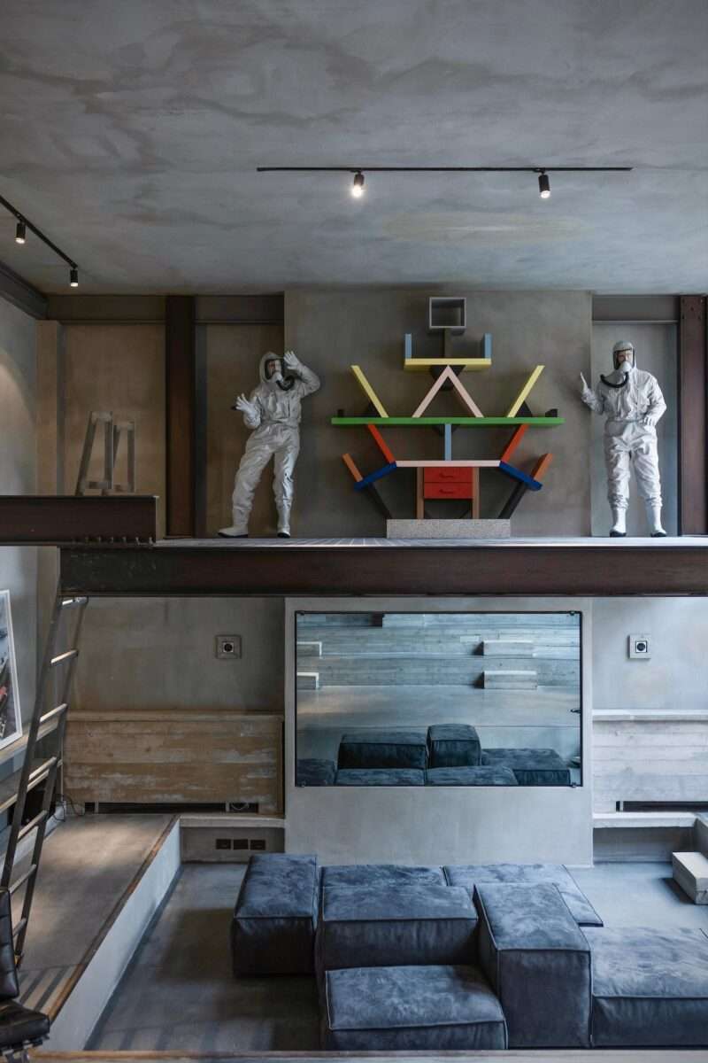 Modern living room with a sunken seating area, grey modular sofas, and a mezzanine featuring colorful geometric shelving and two mannequin figures in white hazmat suits.