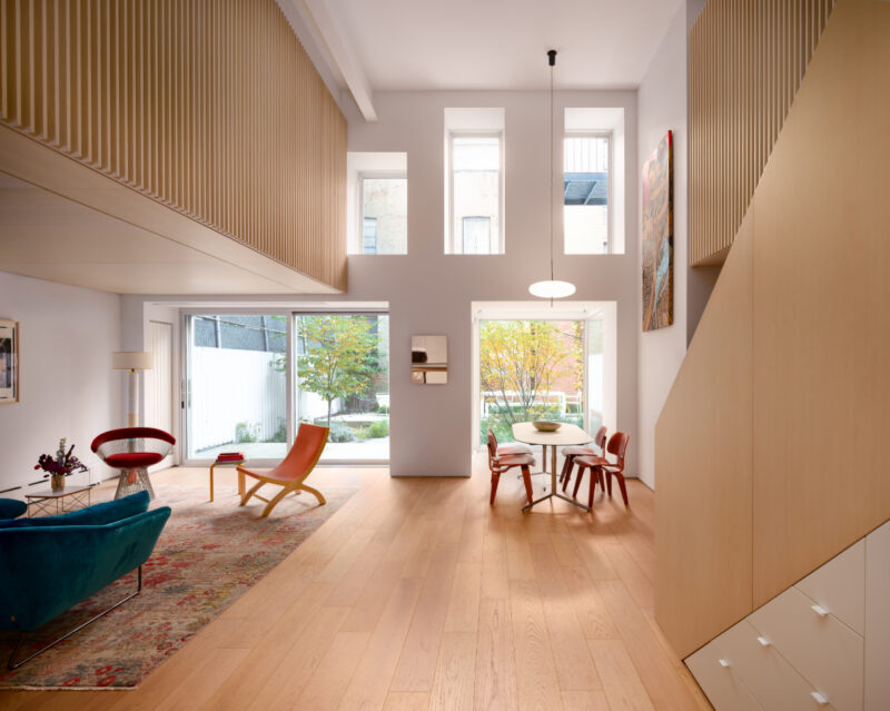 Spacious modern living and dining area with wood floors, high ceilings, tall windows, and minimalist furniture. Large glass doors open to a garden.