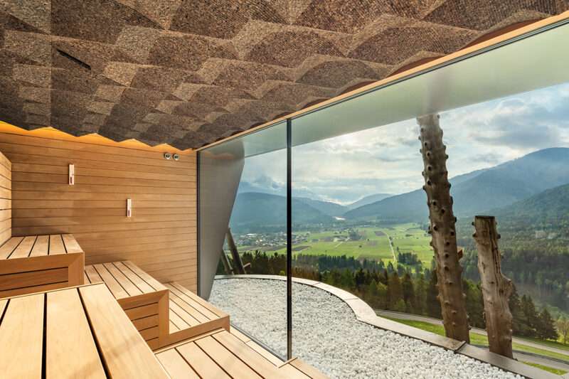 Modern wooden sauna with tiered seating, large floor-to-ceiling window offering a panoramic view of a green valley, mountains, and cloudy sky. White stones decorate the floor near the window, reflecting the refined design seen in the Hotel Hubertus spa expansion.
